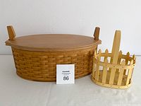 Photo showing large oval basket with wooden handles, fitted wooden lid, and smaller handled basket on the side, both on a white surface.