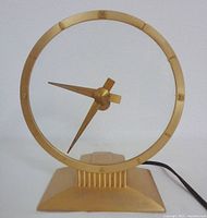 Front view of the Jefferson Golden Hour clock showing the transparent circular glass with gold frame and geometric clock hands on a gold trapezoid base.