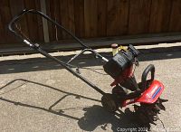 Side view of red Troy-Bilt tiller showing engine, handlebar, and metal tilling blades.