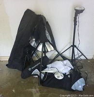 Two light heads with multiple bulb sockets, two soft boxes, two photo umbrellas, light stands, sandbags, and Manfrotto tripod with carrying bag arranged on floor against wall.