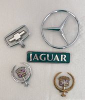 Top view photo of six vintage car hood ornaments and emblems including Chevrolet, Mercedes, Jaguar, and two Cadillac pieces.