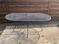 Front view of oval vintage wrought iron outdoor table on concrete, showing mesh surface and three legs with curved supports.