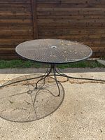 Full view of round wrought iron table with mesh top and umbrella hole, outdoor setting, showing legs and surface condition.