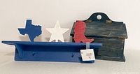 Blue wood shelf with two pegs and three wooden cutouts shaped like Texas, star, and boot atop the shelf against a plain background.