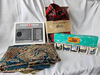 Lot overview showing all 4 main items: boxed set of 5 sake glasses with Japanese art, red plaid heatable booties in box, Swiss+Gear leather wallet in package, and paisley patterned scarf.