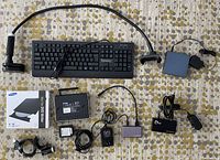 Photo of miscellaneous computer peripherals including wired keyboard, external DVD drive, router, USB hubs, webcam and power supplies on a patterned carpet.