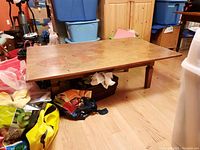 Side angle showing the full wood coffee table placed on wooden floor surrounded by various bags and storage containers. The table has a natural wood finish and low legs.