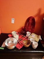 Overview of lot items including red glass vase, hammered metal bowls, mugs, wooden bowls, creamer, sugar container, dish towels, table runner, and red serving plate.
