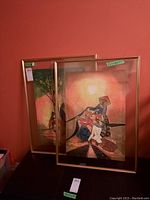 Two framed fabric paintings leaning against a red wall, one featuring abstract figures and the other with figures near trees.