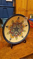 Full view of the 16-inch artisan pottery plate displayed on a black wooden stand on a wooden floor.