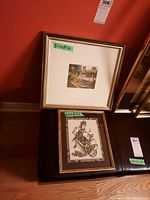 Two framed art prints leaning against wall including a larger silvery framed color print and a smaller gold framed black and white print.