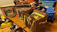 View of 2 Village Grocer collapsible shopping totes with woven wicker pattern, and brown cooler bag with floral design on wooden table among other items.