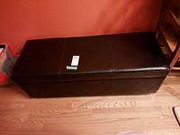Front and top angled view of dark brown leather-looking storage bench showing rectangular shape and stitching detail on lid.