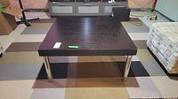 Front view of square coffee table showing laminate top surface and metal legs on multi-colored carpet.