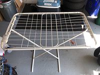 Full view of the metal drying rack showing grid surface and crossed legs, placed on garage floor with visible wear and rust spots.