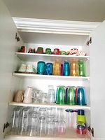 Four shelves of assorted glassware and mugs inside a cabinet including colorful tumblers, ceramic mugs, and clear glasses.