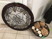 Metal wall art leaning against wall and sea grass bowl filled with decorative balls on the tile floor.