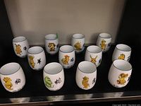 Set of 12 white porcelain cups with colorful cartoon Asian zodiac animal characters and black Chinese characters, displayed together.