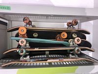 Four skateboards stacked on metal shelf: swirl-wheel board, orange-wheel board, turquoise plastic cruiser board, jester graphic board