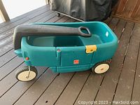 Photo of a green plastic children's wagon with large black handle, white wheels, and red/orange reflectors on sides, showing the exterior side and handle.