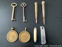 Photo showing two antique-style brass keys, two brass screwdrivers with removable tips, two pen cutting tools with ivory/bone handles, and two commemorative coins with brass holders arranged on a black surface.