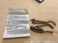 Photo of brass boatswain’s call with attached chain alongside the original instructional history sheet detailing its use and origin.