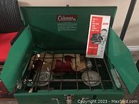 Open view of the Coleman two-burner stove showing green metal body, burners, and protective wind screens.