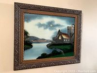 Framed vintage reverse on glass painting showing a lakeside landscape with church and tower under a cloudy sky.