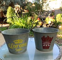 Two metal beer buckets on table in outdoor setting, one with Corona Light logo, other with Weinhard's Red logo