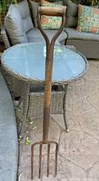 Full view of antique pitchfork showing wooden handle and 4 cast iron tines resting against an outdoor patio table and chairs.