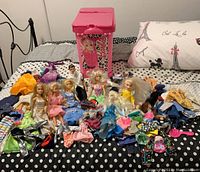 Overview of Barbies, Ken doll, assorted clothes, shoes, and the pink rolling storage case on a bed.