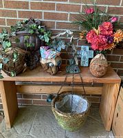 Wide view showing wooden table with three coconut head sculptures, metal wall hangings behind, silk floral arrangements, and hanging wicker basket beneath