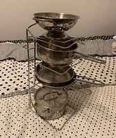 Four stainless steel kids pans stacked vertically on a metal wire storage rack, with a mesh strainer bowl at the top