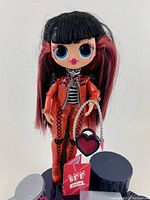 Front view of LOL OMG Surprise Spicy Babe doll with black and pink hair, orange clothing, holding a heart-shaped handbag, and red booklet on a white background.