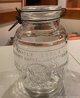 Front angle view showing embossed brand and emblem on clear glass jar, metal latch and glass lid visible.