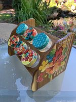 Image showing angled top view of wooden spice rack holding 6 glass jars with colorful floral and geometric lids; floral painted side panel visible