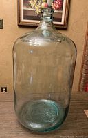 Full front view of a large green-tinted glass carboy jug standing on a table near a painting and electrical outlet.