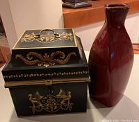 Front view of the black metal chest showing gold gryphon designs and the rustic red vase beside it on a table.