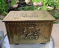 Front and side view of the coal or kindling box showing embossed brass panel depicting a tavern scene with people playing music and socializing outdoors by trees and a brick building.