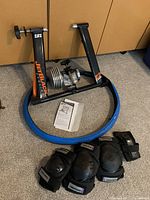 Overall view of the bike trainer stand, blue tire cover, instruction manual, and three black knee pads on carpeted floor.
