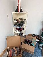 Corner white shelf with stacked books including art and nature books. Three open cardboard boxes on floor with more books inside.
