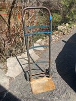 Front view of metal frame hand truck dolly with solid rubber wheels and wooden base plate on asphalt driveway.