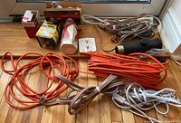 Photo showing multiple orange and brown extension cords, power bar, light bulbs in box packaging, three-prong plug adapter, and black torch/flashlight on hardwood floor.