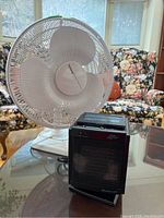 Holmes electric fan and Duracraft portable heater on glass table with floral couch background.