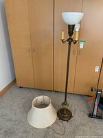 Full view of vintage floor lamp standing next to a wooden wardrobe, with beige fabric shade on floor beside lamp base.