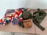 Top-down view showing Union Jack flag, steel helmet, binoculars, leather case, green supply bag, and military belt.