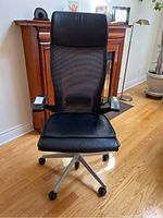 Front view of Haworth highback office chair showing black leather seat, mesh backrest, armrests with adjustment controls, and silver base with five caster wheels.