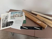 Overview of two prints lying flat on a table, black parallel ruler, signed letter, and four rolled art posters in tubes.