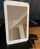 Front view of Samsung Galaxy Tab3 Lite tablet standing upright with charger next to it on a reflective surface.