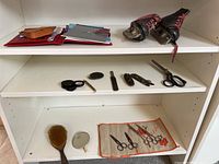 Overview of white shelving containing various items: vintage children's roller skates, employee records or folders, multiple scissors and small tools, hair brush, hand mirror, and embroidered cloth with scissors.